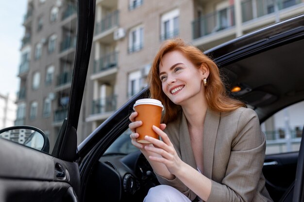 Shot van een volwassen vrouw die in haar auto zit met een open deur en koffie drinkt uit een reismok