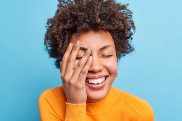 shot van gelukkige krullende jonge Afro-Amerikaanse vrouw sluit ogen en grijnst van vreugde houdt handpalm op gezicht drukt authentieke emoties uit geïsoleerd over blauwe muur heeft plezier natuurlijke frisse schone huid
