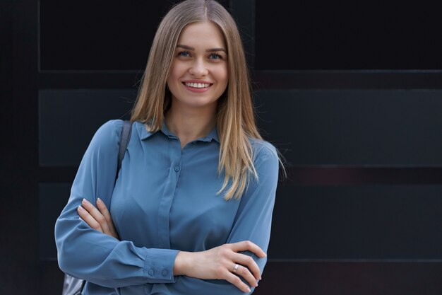 Shot van mooie jonge zakenvrouw blauw chiffon overhemd dragen terwijl met gevouwen armen op zwarte muur