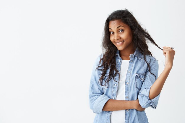 shot van schattige donkere vrouw die speelt met haar lange golvende haar, stralend gezicht en zachte glimlach die haar schoonheid verheugt. De vrolijke vrouw kleedde zich terloops hebbend en flirtende uitdrukking.