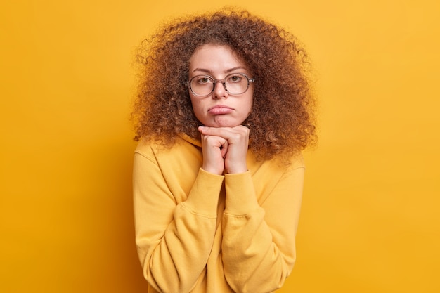 shot van teleurgestelde verdrietige Europese vrouw met krullend haar voelt zich overstuur houdt handen onder kin portemonnees lippen gekleed in sweatshirt geïsoleerd over gele muur voelt lastig en verveeld