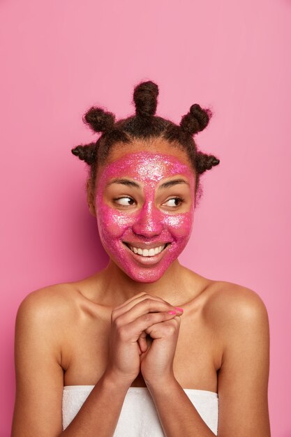 shot van vrolijke schoonheid model staat gewikkeld in een handdoek, past voedend masker toe, houdt de handen tegen elkaar gedrukt, kijkt opzij, poseert over roze muur. Gezichtsverzorging, wellness, cosmetologie concept