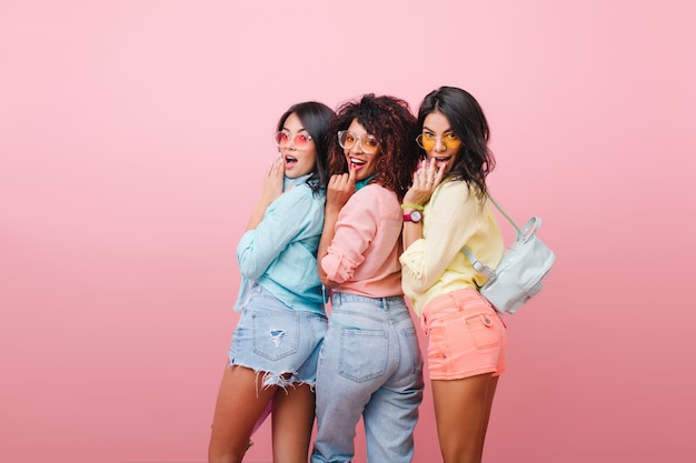 Sierlijk Europees meisje in blauw shirt kijkt over de schouder, staande in de buurt van internationale vriendinnen. Binnenfoto van drie charmante dames in zomerkleren die voor de gek houden.