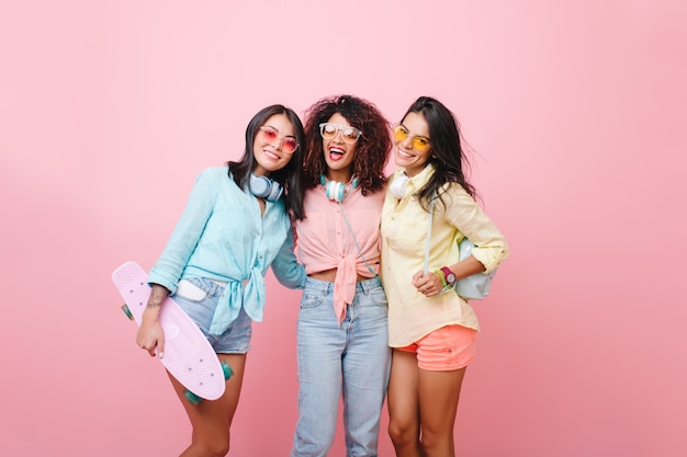 Sierlijk meisje in geel shirt met lederen rugzak poseren in de buurt van afrikaanse krullende vriend in spijkerbroek. vrolijke zwarte jonge vrouw in zonnebril die zich tussen europese en latino dames bevindt.