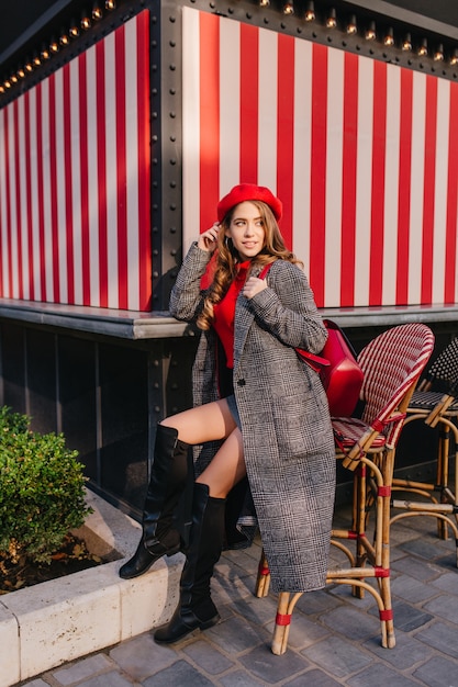 Gratis foto sierlijk meisje in lange tweed jas zittend op een stoel op straat en iemand te wachten