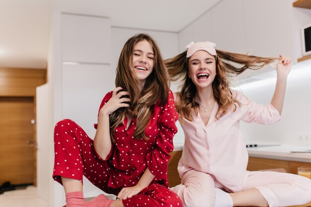 Sierlijk wit meisje met krullend kapsel genieten van Goedemorgen met zus. Binnenopname van geïnspireerde blanke vrouwelijke modellen in nachtkostuums die met hun haar spelen.
