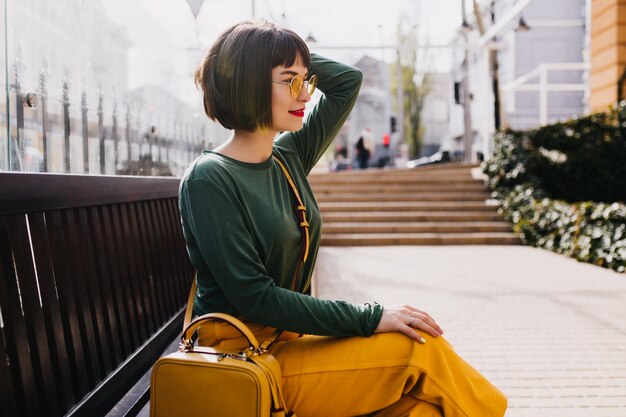 Sierlijke kortharige meisje in zonnebril buiten ontspannen. Schattige vrouw in groene trui poseren op bankje in zonnige dag.