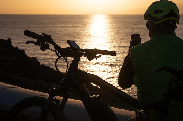Silhouet van man met behulp van mobiele telefoon bij zonsondergang met helm na het fietsen langs de zee