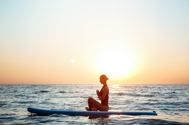 Silhouet van mooie vrouw het beoefenen van yoga op surfplank bij zonsopgang.