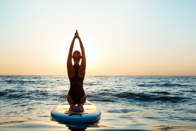 Silhouet van mooie vrouw het beoefenen van yoga op surfplank bij zonsopgang.