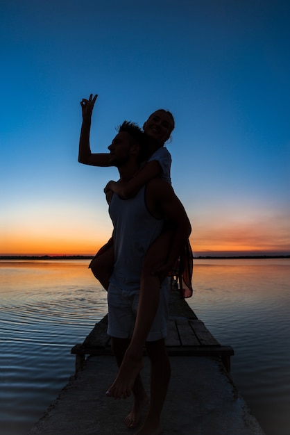 Silhouetten van het jonge mooie paar rusten die zich verheugen bij zonsopgang dichtbij meer