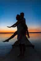 Gratis foto silhouetten van het jonge mooie paar rusten die zich verheugen bij zonsopgang dichtbij meer