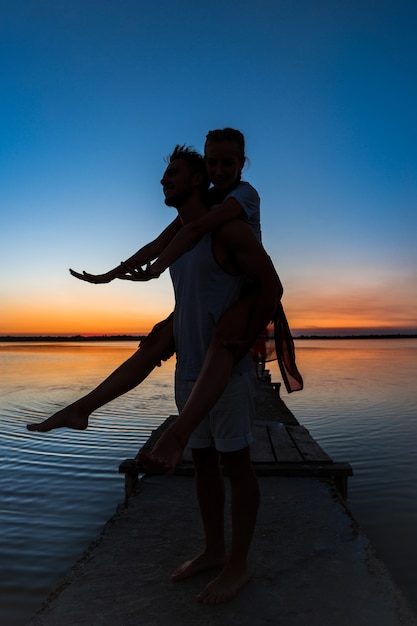 Silhouetten van het jonge mooie paar rusten die zich verheugen bij zonsopgang dichtbij meer