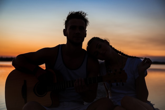 Gratis foto silhouetten van het jonge mooie paar rusten die zich verheugen bij zonsopgang dichtbij meer