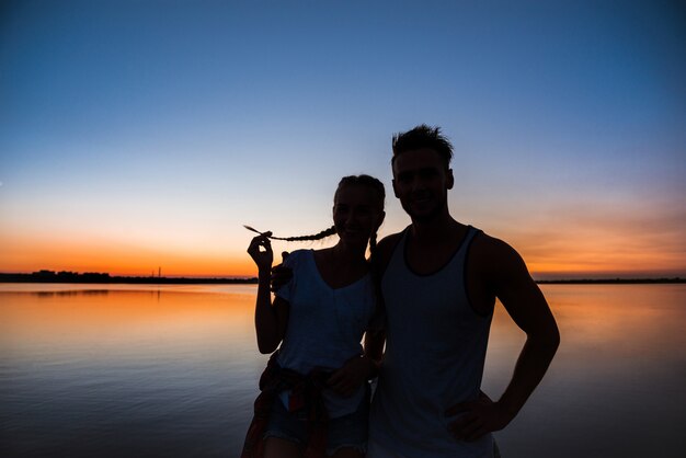 Silhouetten van het jonge mooie paar rusten die zich verheugen bij zonsopgang dichtbij meer