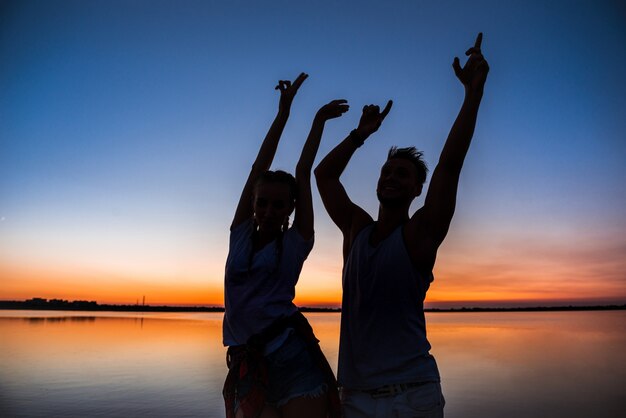 Silhouetten van het jonge mooie paar rusten die zich verheugen bij zonsopgang dichtbij meer