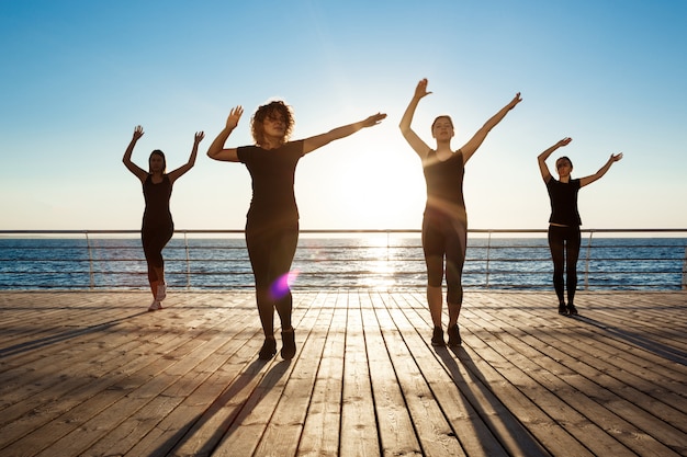 Gratis foto silhouetten van sportieve vrouwen die zumba dichtbij overzees dansen bij zonsopgang