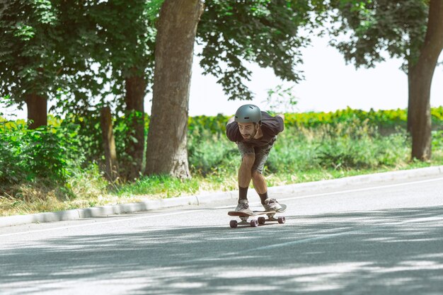 Skateboarder doet een truc in de buurt van weide in zonnige dag. Jonge man in uitrusting rijden en longboarden op het asfalt in actie. Concept van vrijetijdsbesteding, sport, extreem, hobby en beweging.