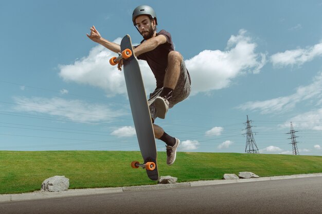 Skateboarder doet een truc in de straat van de stad op zonnige dag. Jonge man in uitrusting rijden en longboarden in de buurt van weide in actie. Concept van vrijetijdsbesteding, sport, extreem, hobby en beweging.