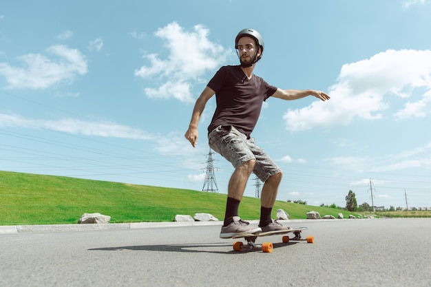 Skateboarder doet een truc in de straat van de stad op zonnige dag. Jonge man in uitrusting rijden en longboarden in de buurt van weide in actie. Concept van vrijetijdsbesteding, sport, extreem, hobby en beweging.