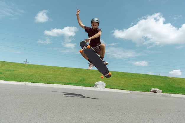 Skateboarder doet een truc in de straat van de stad op zonnige dag. jonge man in uitrusting rijden en longboarden in de buurt van weide in actie. concept van vrijetijdsbesteding, sport, extreem, hobby en beweging.