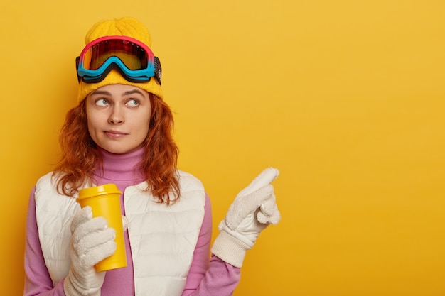 Skiër met rood haar, draagt gele hoed, wijst op een lege plek, houdt afhaalkoffie vast, demonstreert winterlandschap, staat tegen gele achtergrond