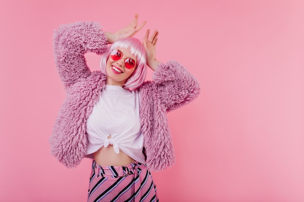 Slank mooi meisje in pluizig jasje gek rond op roze muur. Geïnteresseerde jonge vrouw in stijlvolle peruke uiting van geluk tijdens fotoshoot