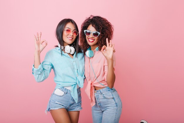 slanke Amerikaanse dames in zomerkleding poseren met een blije glimlach. Betoverende blanke vrouw met glanzend kapsel genieten van vrije tijd met Afrikaanse vriendin.