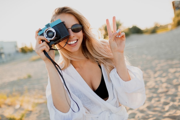 Slanke blonde gelukkige vrouw die retro camera houdt en pret op warm zonnig strand heeft. Zomervakantie en reizen concept. Natuurlijke schoonheid, vakantie in Azië. Trendy zonnebril, witte outfit.