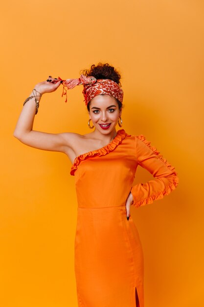 Slanke dame in elegante oranje jurk en hoofdband in Afrikaanse stijl glimlacht lief tegen de oranje ruimte.