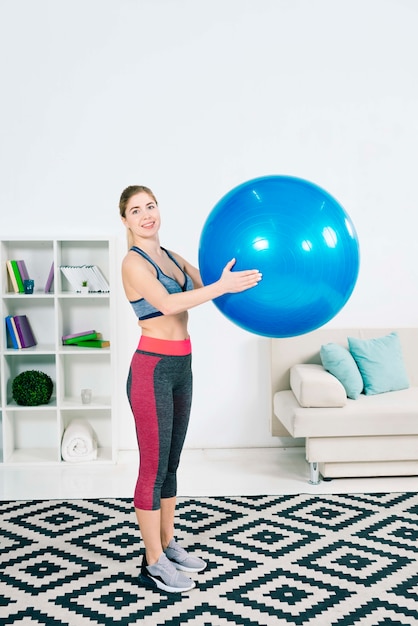 Slanke geschikte jonge vrouw die zich in de woonkamer bevindt die blauwe pilatesbal houdt