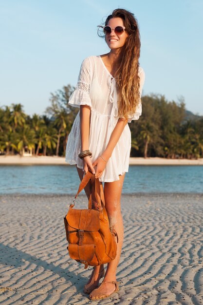 Slanke mooie vrouw in witte katoenen jurk wandelen op tropisch strand op zonsondergang lederen rugzak te houden.