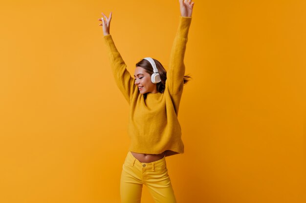 Slanke positieve vrouw in gele broek dansen met omhoog handen. Indoor portret van zalige meisje in grote koptelefoon genieten van goed lied.