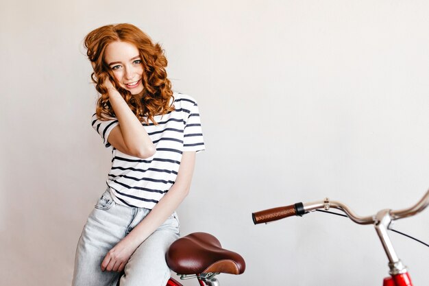 Slanke schattige dame in spijkerbroek poseren met fiets. Binnenfoto van blij krullende vrouw die zich dichtbij fiets bevindt en positieve emoties uitdrukt.