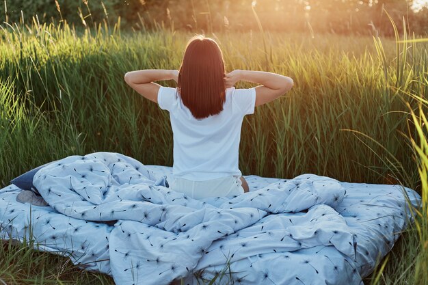 Slanke vrouw met donker haar met een wit casual t-shirt dat achterstevoren poseert met opgeheven armen, handen strekkend na het slapen, genietend van zonsopgang in groene weide in prachtige natuur.