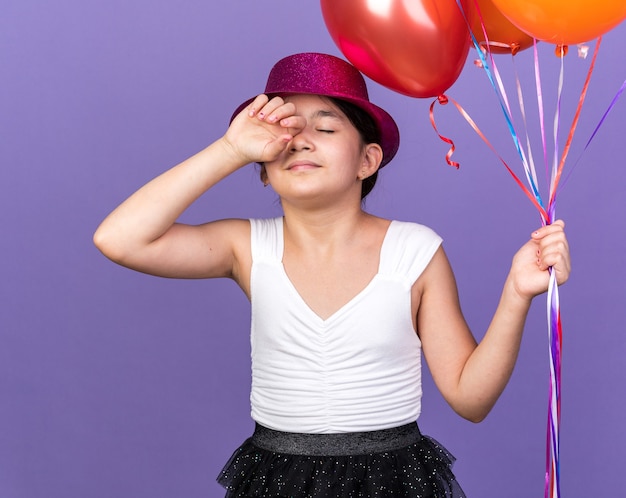 slaperig jong kaukasisch meisje met violet feestmuts met heliumballonnen geïsoleerd op paarse muur met kopieerruimte