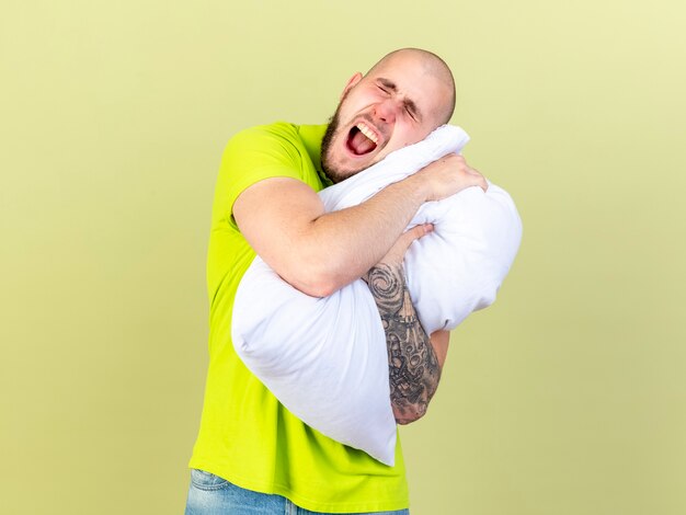 Slaperige jonge zieke man gaapt met gesloten ogen en legt hoofd op kussen geïsoleerd op olijfgroene muur