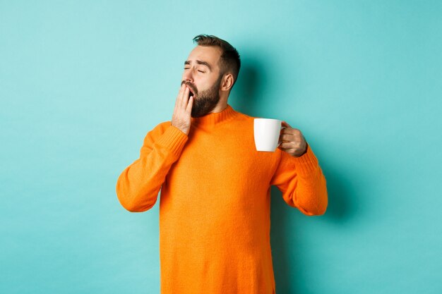 Slaperige knappe man koffie drinken en geeuwen, staande in oranje trui tegen licht turkooizen muur