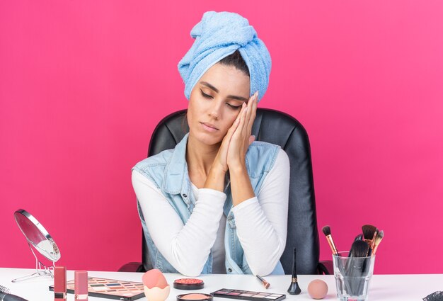Slaperige, mooie blanke vrouw met gewikkeld haar in een handdoek zittend aan tafel met make-uptools die het hoofd op haar handen leggen