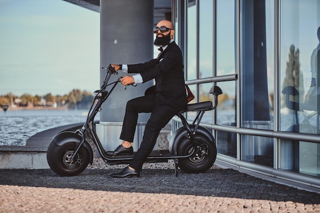 Slimme aantrekkelijke zakenman zit op zijn electro scooter terwijl hij poseert voor fotograaf.