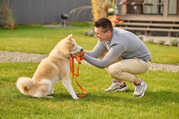 Slimme hond. Zijwaarts naar de camera van een vrolijke jonge volwassen man die op herfstdag in de buurt van een landhuis gehurkt een vriendelijke en slimme hond aanraakt met een riem