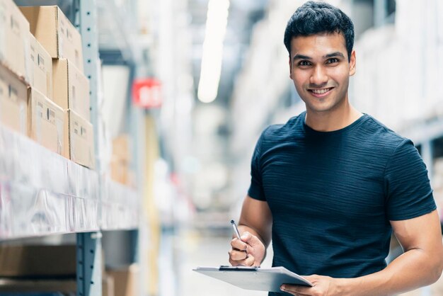 Slimme Indiase ingenieur met een veiligheidshelm die de voorraadcontrole en het productbeheer van de kartonvoorraad doet op de achtergrond van het fabrieksmagazijn