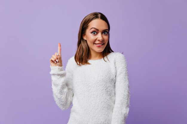 Slimme meid van 23 jaar oud die op haar lip bijt van vreugde bij het ontstaan van een nieuw briljant idee. Portret van emotionele vrouw in comfortabele kleding tegen paarse muur