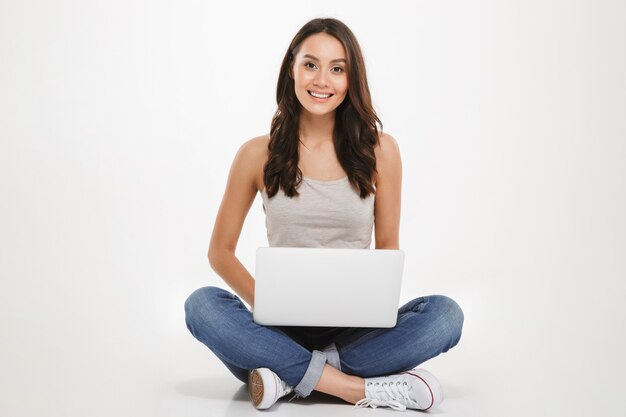 slimme vrouw met lang bruin haar zittend met benen gekruist op de vloer met behulp van zilveren laptop en kijken op camera, geïsoleerd over witte muur