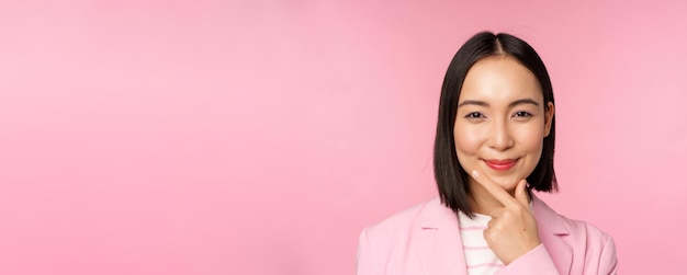 Sluit het portret van een glimlachende Aziatische werkende dame in een pakzakenvrouw die bedachtzaam nadenkt of beslist over een roze achtergrond