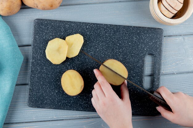 Sluit mening die van vrouwenhanden aardappel met mes op scherpe raad op houten achtergrond omhoog snijden