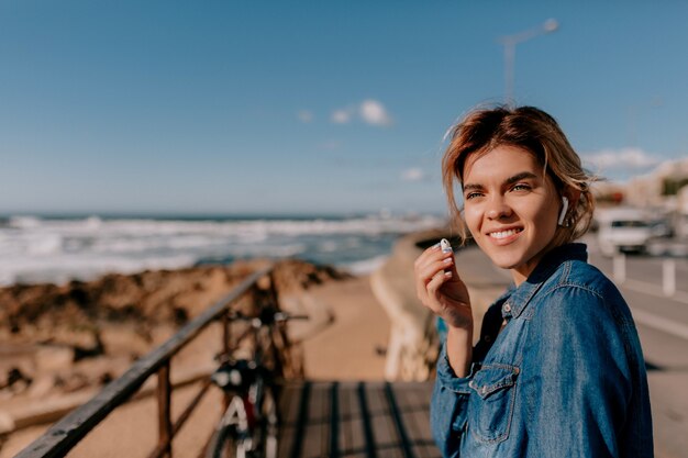 Sluit omhoog buiten portret van mooie glimlachende vrouw die denimoverhemd draagt dat airpods houdt en op de oceaan in zonnige dag kijkt