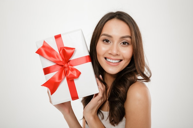 Sluit omhoog foto die van vrolijke vrouw gift-verpakte doos met rode boog op camera tonen die geluk en verrukking uitdrukken, die over wit wordt geïsoleerd