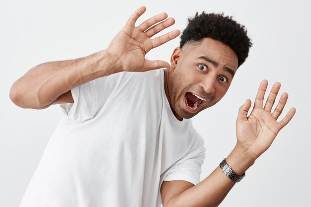 Sluit omhoog geïsoleerd portret van grappige jonge god-kijkende aantrekkelijke tan-gevilde mens met afrokapsel in het modieuze witte t-shirt gillen, bangend door vreemdeling op straat.