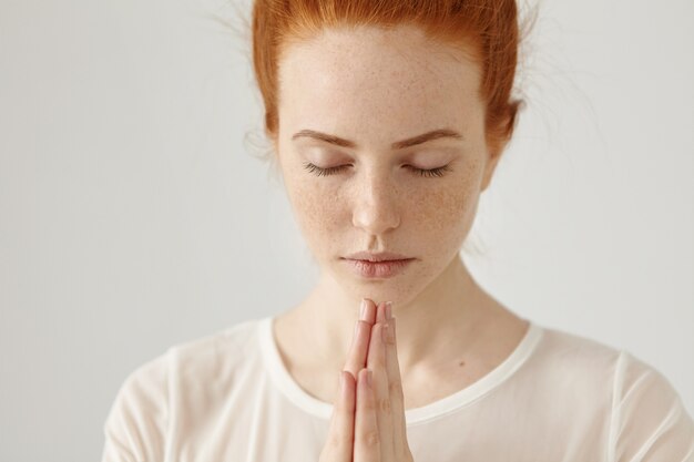 Sluit omhoog geschoten van godsdienstige jonge gembervrouw in witte blouse die mediteren of bidden houdend gesloten ogen en samen gedrukte handen, hopend op het beste. Mensen, religie, spiritualiteit, gebed